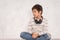 Studio shot of a young boy wearing a white shirt and jeans with headphones siting on the floor by the wall and thinks or sad about