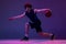 Studio shot of young muscular basketball street player dribbling ball isolated on purple background in neon. Concept of