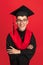 Studio shot of young smiling, cheerful student, graduate in mortarboard isolated over red studio background.
