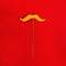 Stuffed yellow moustache toy on a stick, red background, flat lay.
