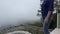 Stunned female traveler standing at green abyss in the mountain watching white cloud fog coming closer. Elderly woman