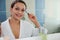 Stunning multi-ethnic woman in white bathrobe reflecting in bathroom mirror, smiling while applying mascara on eyelashes