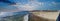 A stunning panoramic shot of large waves form the vast blue ocean rolling into the beach filled with people relaxing