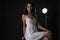 Stunning portrait of a sexy young brunette girl in a white dress sitting on a high chair on a black background. Low key. Beauty