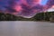 A stunning shot of the vast still waters of a lake surrounded by lush green trees with powerful red clouds at sunset