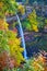 Stunning tall waterfall over cliffs is surrounded by colorful peak fall foliage