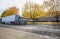 Stylish big rig gray semi truck with semi trailer standing on the autumn road with yellow trees at industrial area waiting for