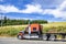 Stylish black and orange big rig semi truck transporting empty flat bed semi trailer driving on the road along the summer meadow