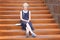 Stylish blonde preteen girl in a dress sits on the stairs with decorative rust in a modern building