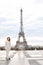 Stylish blonde woman standing near Eiffel Tower in white overalls.