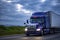 Stylish blue big rig semi truck transporting frozen cargo in refrigerator semi trailer running on the evening wide highway road
