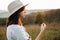 Stylish boho girl holding dried herb in sunny light at atmospheric sunset in meadow. Calm hipster woman in poncho and hat enjoying