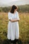 Stylish boho girl walking in grass and wildflowers in sunny meadow at  atmospheric sunset. Happy hipster woman in linen rustic