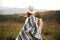 Stylish boho girl walking in sunny light at atmospheric sunset in meadow. Happy hipster woman in poncho and hat enjoying traveling