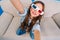 Stylish brightful selfie portrait of joyful little girl in 3d glasses with long brunette hair smiling to camera on couch