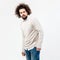 Stylish brown-haired curly guy with a beard dressed in beige jumper over a white shirt and jeans poses in the studio on