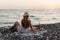 Stylish and carefree woman wearing a straw hat is having a serene picnic on a pebble beach during a sunset