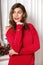 Stylish charming girl with red lipstick dressed in a red sweater poses poses against a white wall in the room