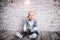 A stylish child, a boy, one year old, sit on a wooden floor and on a brick wall background. He is dressed in a junky jacket, junks