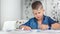 Stylish confident kid boy drawing paper image on desk surrounded by classroom white interior