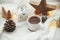 Stylish cup of tea with modern christmas houses, pine cone, wooden star and tree decor, golden lights on warm blanket on