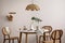 The stylish dining room with round table, rattan chair, lamp and kitchen accessories. Green leaf in vase. Beige wall. Home decor