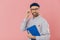 Stylish distant worker wears transparent glasses, stylish hat, shirt, holds blue textbook, uses pencils for writing information,