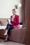 Stylish elegant businesswoman wearing red jacket sitting on bed in hotel