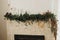 Stylish fireplace mantel decorated with christmas branches, bells garland, wooden ornaments and house decoration. Festive rustic
