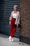 Stylish full length woman standing near wall dressed in sweater, shirt, red pants, white leather boots with heels