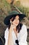 Stylish girl in fashionable look sitting on wooden stairs on beach cliff at sea in sunny light. Happy young boho woman in hat