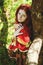 Stylish girl with freckles and red hair in red long dress holding red poppy flower at green field on nature