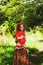 Stylish girl with freckles and red hair in red long dress holding red poppy flower at green field on nature