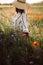 Stylish girl in hat walking in wildflowers in sunset light in summer meadow. Young woman in linen dress walking among poppy and