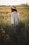 Stylish girl in hat walking in wildflowers in sunset light in summer meadow. Young woman in linen dress walking among poppy and