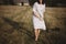 Stylish girl in linen dress and hat running barefoot in grass in sunny field at village. Boho woman relaxing in countryside,