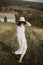 Stylish girl in linen dress and hat running barefoot in grass in sunny field at village. Boho woman relaxing in countryside,