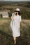 Stylish girl in linen dress and hat walking barefoot in grass in sunny field at village. Boho woman relaxing in countryside,