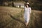 Stylish girl in linen dress and hat walking barefoot in grass in sunny field at village. Boho woman relaxing in countryside,