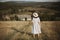 Stylish girl in linen dress and hat walking barefoot in grass in sunny field at village. Boho woman relaxing in countryside,