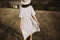 Stylish girl in linen dress and hat walking barefoot among herbs and wildflowers in sunny field in mountains. Boho woman relaxing