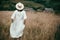 Stylish girl in linen dress and hat walking among herbs and wildflowers in field. Boho woman enjoying day in countryside, simple