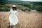 Stylish girl in linen dress and hat walking among herbs and wildflowers in field. Boho woman enjoying day in countryside, simple