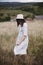 Stylish girl in linen dress and hat walking among herbs and wildflowers in field. Boho woman relaxing in countryside, simple slow
