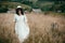 Stylish girl in linen dress and hat walking among herbs and wildflowers in field. Boho woman smiling and relaxing in countryside,