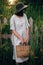 Stylish girl in linen dress holding rustic straw basket at wooden fence among green cane. Boho woman in hat relaxing and posing in