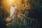 Stylish girl in linen dress holding rustic straw basket at wooden fence  in sunset light. Boho woman relaxing and posing in summer