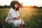 Stylish girl in linen dress smelling poppy flower in meadow in sunset light with flowers in rustic straw basket. Boho woman in hat