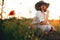 Stylish girl in linen dress smelling poppy flower in meadow in sunset light with flowers in rustic straw basket. Boho woman in hat