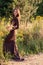 Stylish girl in long dress with rustic bouquet walking in meadow in sunset